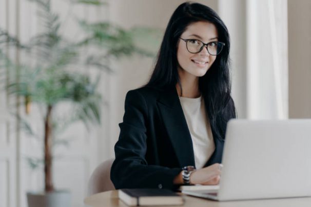 Smiling well dressed young woman secretary with dark hair, works on laptop computer at desktop, makes project or research work. Sales manager in modern office, cozy workplace. Remote job concept